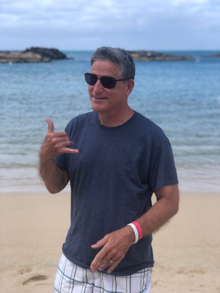 Andy Carignan on a beach giving a traditional Hawaiian Shaka sign